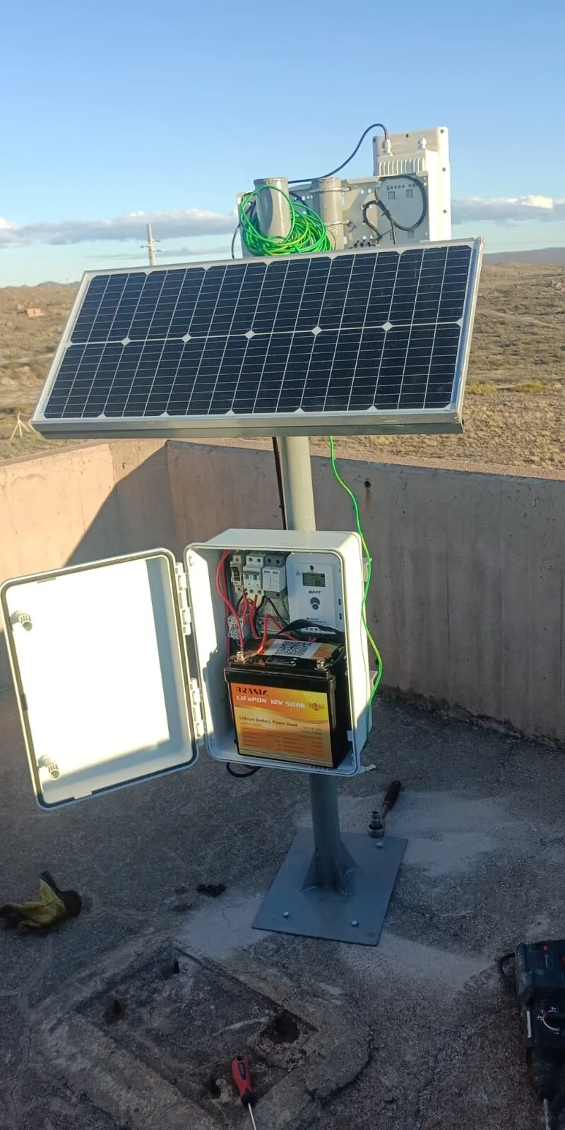Estación remota con panel solar, gabinete abierto con batería y antenas sobre un pedestal de acero
