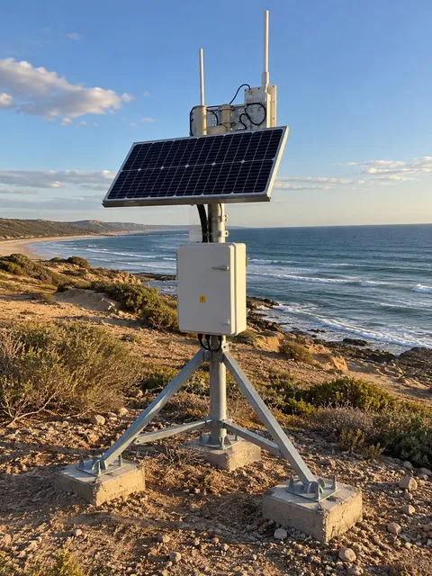 Ilustración de una estación solar de monitoreo sobre un acantilado costero, con panel solar, gabinete y antenas